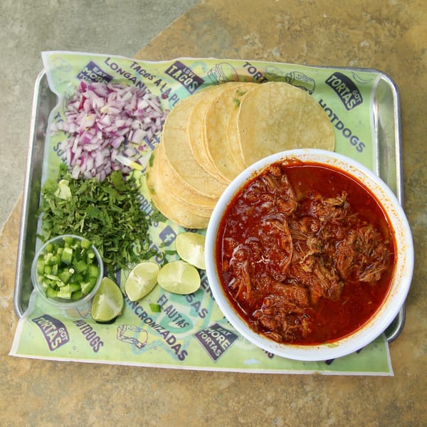 Birria tacos kit with shredded meat in red sauce, tortillas, cilantro, onions, jalapeños, and limes.