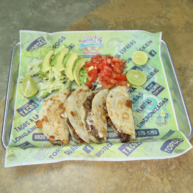 Four El Vagoncito Chilango tacos with shredded lettuce, avocado slices, diced tomatoes, and lime wedges.