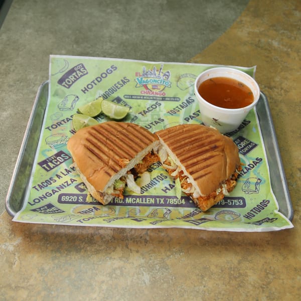 A Mexican torta, cut in half, with limes and a cup of consommé on a branded paper liner.