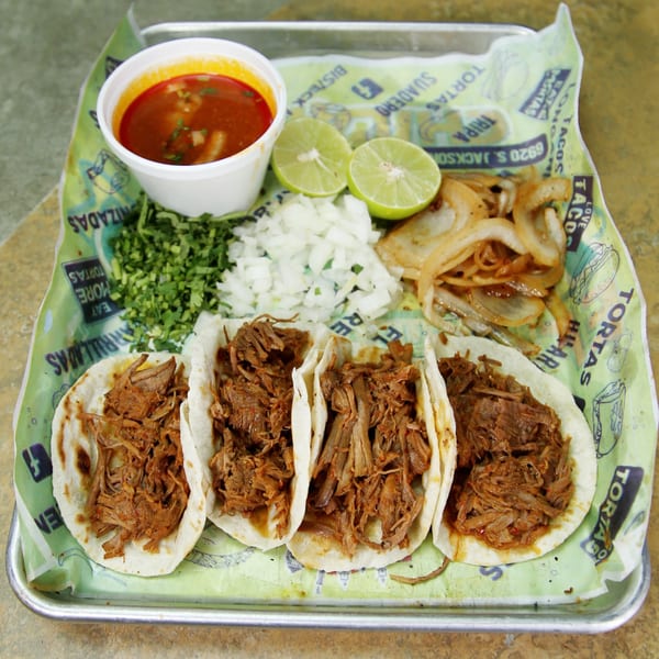Four El Vagoncito Chilango birria tacos with sides of broth, limes, cilantro, onions, and grilled onions.