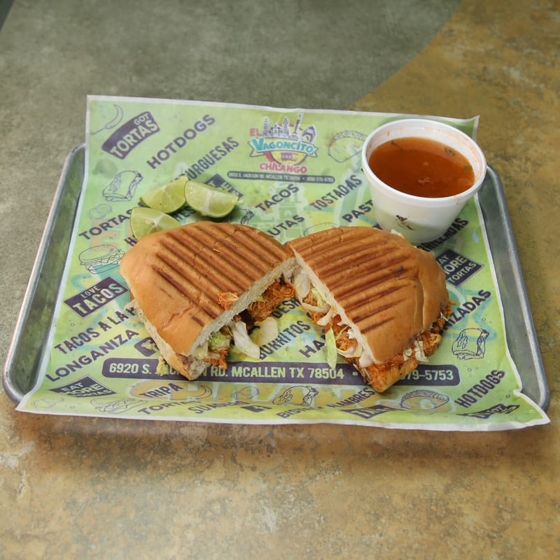 A Mexican torta, cut in half, with limes and a cup of broth on a branded paper-lined tray.