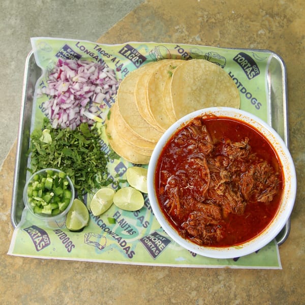 A tray of birria tacos with a bowl of stew, tortillas, cilantro, onions, jalapeños, and limes.