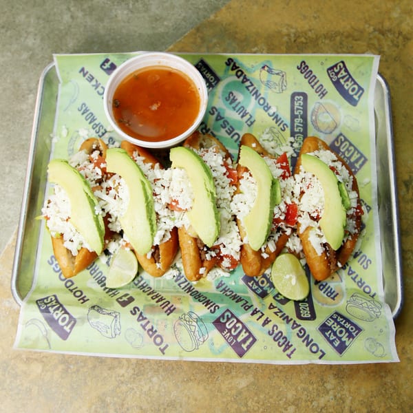 Six El Vagoncito Chilango hot dogs topped with avocado, cheese, and salsa on a tray.