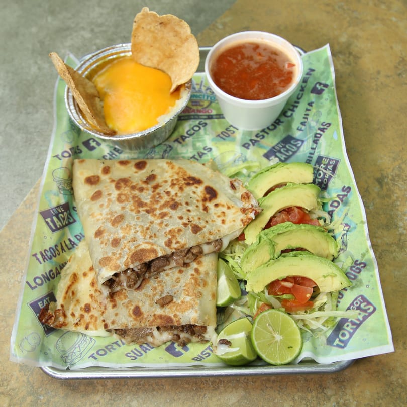 A quesadilla with meat and cheese, served with avocado, limes, salsa, and nachos with cheese.