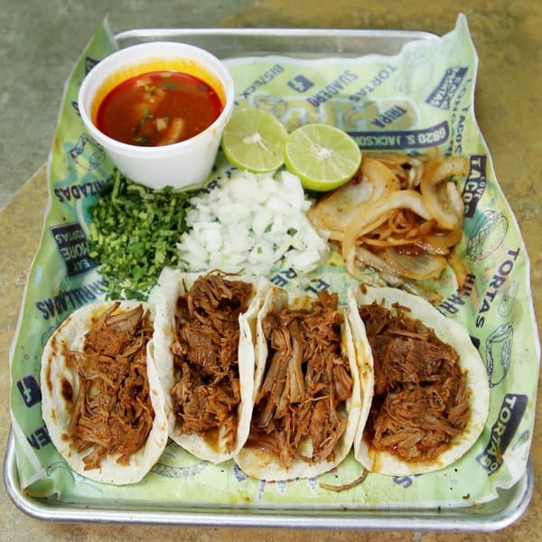 Four El Vagoncito Chilango tacos with shredded meat, a side of broth, limes, onions, and cilantro.