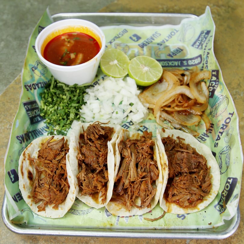 Four El Vagoncito Chilango tacos with shredded meat, a side of broth, limes, onions, and cilantro.