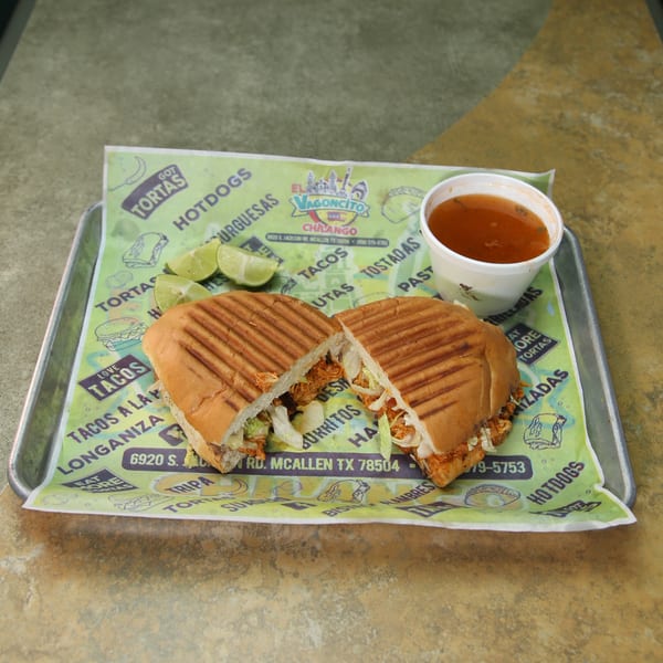 A Mexican torta, cut in half, with limes and a cup of consommé on a branded paper liner.