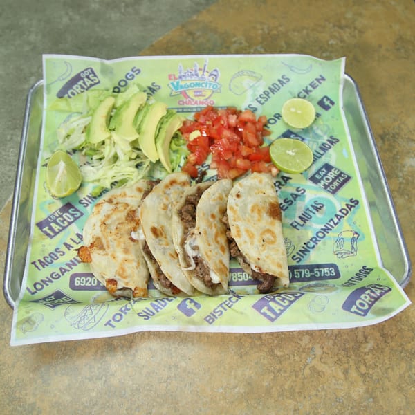 Four El Vagoncito Chilango tacos with avocado, diced tomatoes, and lime wedges on a branded paper.