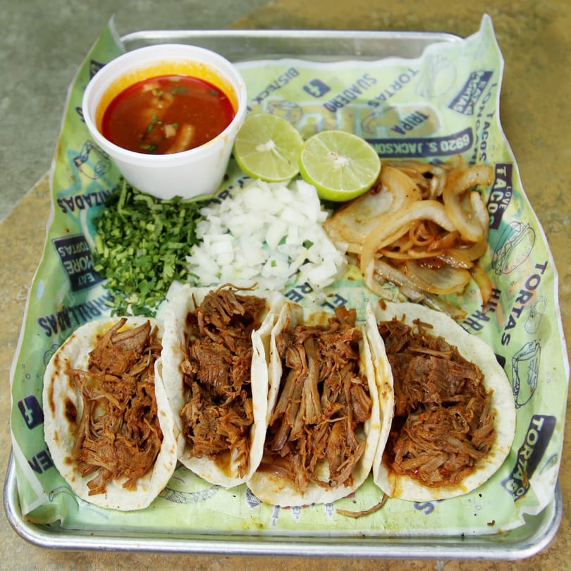 Four El Vagoncito Chilango birria tacos with a side of consommé, limes, cilantro, onions, and grilled onions.